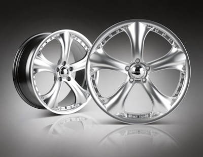 A pair of sleek, metallic alloy wheels with a shiny, polished finish is set against a gradient background. The wheels feature a five-spoke design, and the surface reflects light, emphasizing their smooth and modern appearance.