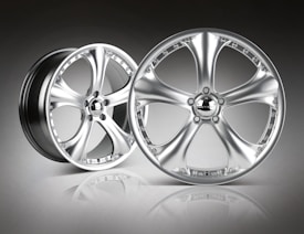 A pair of sleek, metallic alloy wheels with a shiny, polished finish is set against a gradient background. The wheels feature a five-spoke design, and the surface reflects light, emphasizing their smooth and modern appearance.