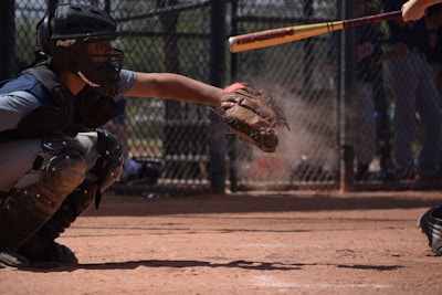 A baseball player in catcher gear is crouching down, ready to catch a baseball. Another player is in the process of swinging a bat. Dust rises from the ground, indicating the intensity of the play. A chain-link fence surrounds the field, with players and spectators visible in the background.