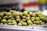 A pile of green olives is arranged on a metallic tray, with a blurred background indicating a market or deli setting. The olives appear fresh and are of various shades of green.