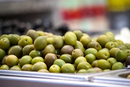 A pile of green olives is arranged on a metallic tray, with a blurred background indicating a market or deli setting. The olives appear fresh and are of various shades of green.