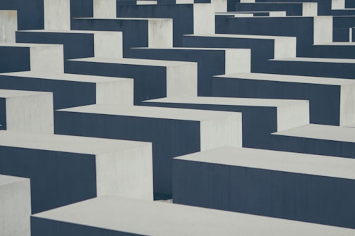 Concrete slabs are arranged in a grid pattern, creating a maze-like appearance with alternating dark and light surfaces. The slabs have varying heights, casting shadows and enhancing the geometric design.