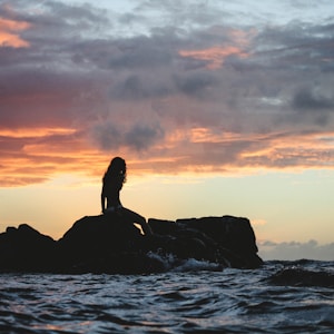 silhouette photo of woman sitting on rock during golden hour