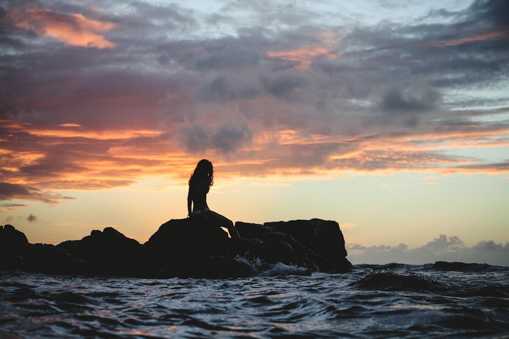 silhouette photo of woman sitting on rock during golden hour