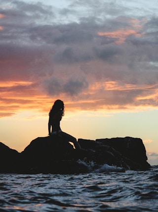 silhouette photo of woman sitting on rock during golden hour