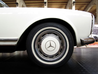 A close-up view of a vintage white car's rear wheel, featuring a classic hubcap with a prominent Mercedes-Benz logo. The tire is mounted on a clean, polished wheel, and the smooth paint of the car body curves gracefully above it. The car is positioned indoors, with industrial-style beams visible in the background.