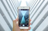 A hand holds a smartphone displaying a symmetrical architectural view with tall, converging walls creating a tunnel-like effect. The alignment and the perspective captured on the phone's screen mirror the visual scene in the background.