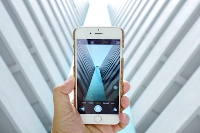 A hand holds a smartphone displaying a symmetrical architectural view with tall, converging walls creating a tunnel-like effect. The alignment and the perspective captured on the phone's screen mirror the visual scene in the background.