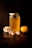 A jar filled with golden honey sits on a dark surface, capped with a lid featuring a honeycomb pattern. Beside it are fresh mandarin segments and a whole mandarin, accompanied by delicate white flowers with hints of peach color. The scene is lit with warm, subdued lighting that highlights the rich amber tones of the honey and the bright orange of the fruit.