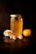 A jar filled with golden honey sits on a dark surface, capped with a lid featuring a honeycomb pattern. Beside it are fresh mandarin segments and a whole mandarin, accompanied by delicate white flowers with hints of peach color. The scene is lit with warm, subdued lighting that highlights the rich amber tones of the honey and the bright orange of the fruit.