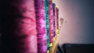 Multiple spools of thread are arranged in a row, each spool featuring a different color. The threads range from shades of pink, green, and purple, displayed on a wooden surface with a blurred background.