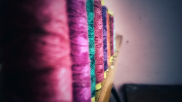 Multiple spools of thread are arranged in a row, each spool featuring a different color. The threads range from shades of pink, green, and purple, displayed on a wooden surface with a blurred background.