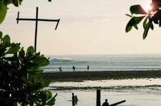 A serene beach scene showing people walking along the shoreline. The foreground is framed by green leaves and branches, while a cross-shaped structure stands prominently to the left. The ocean appears calm, with gentle waves breaking in the distance.