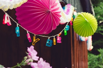 Colorful paper lanterns and letters spelling out 'HAPPY BIRTHDAY' hang from a string. Bright pink and green fans stand out against a wooden background. Below, pink flowers are partially visible, adding a natural touch to the festive decor.