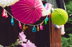 Colorful paper lanterns and letters spelling out 'HAPPY BIRTHDAY' hang from a string. Bright pink and green fans stand out against a wooden background. Below, pink flowers are partially visible, adding a natural touch to the festive decor.