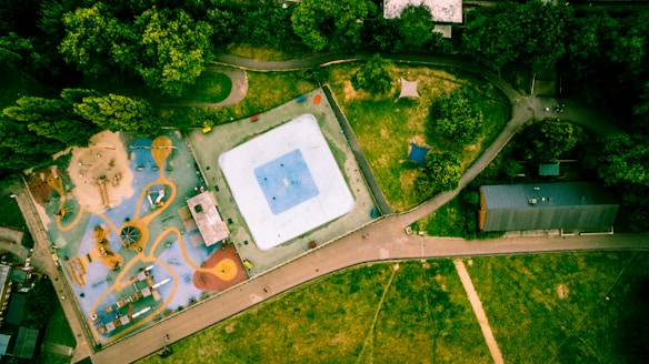 An aerial view of a park showcasing a playground with colorful structures and an adjacent open grass area. The playground is equipped with slides, climbing frames, and a sandpit. There are pathways and a building nearby, surrounded by lush green trees.
