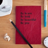 A red textured notebook is centered on a wooden table with the words 'Be brave, Be bold, Be beautiful, Be you' written in black text. Beside the notebook, there is a pencil with sharpened wood shavings nearby. A cup of coffee with a spoon rests on a saucer to the upper left, and a blue and gold pen lies on the right side. The edge of a wristwatch is visible at the bottom right corner.