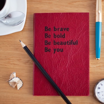 A red textured notebook is centered on a wooden table with the words 'Be brave, Be bold, Be beautiful, Be you' written in black text. Beside the notebook, there is a pencil with sharpened wood shavings nearby. A cup of coffee with a spoon rests on a saucer to the upper left, and a blue and gold pen lies on the right side. The edge of a wristwatch is visible at the bottom right corner.