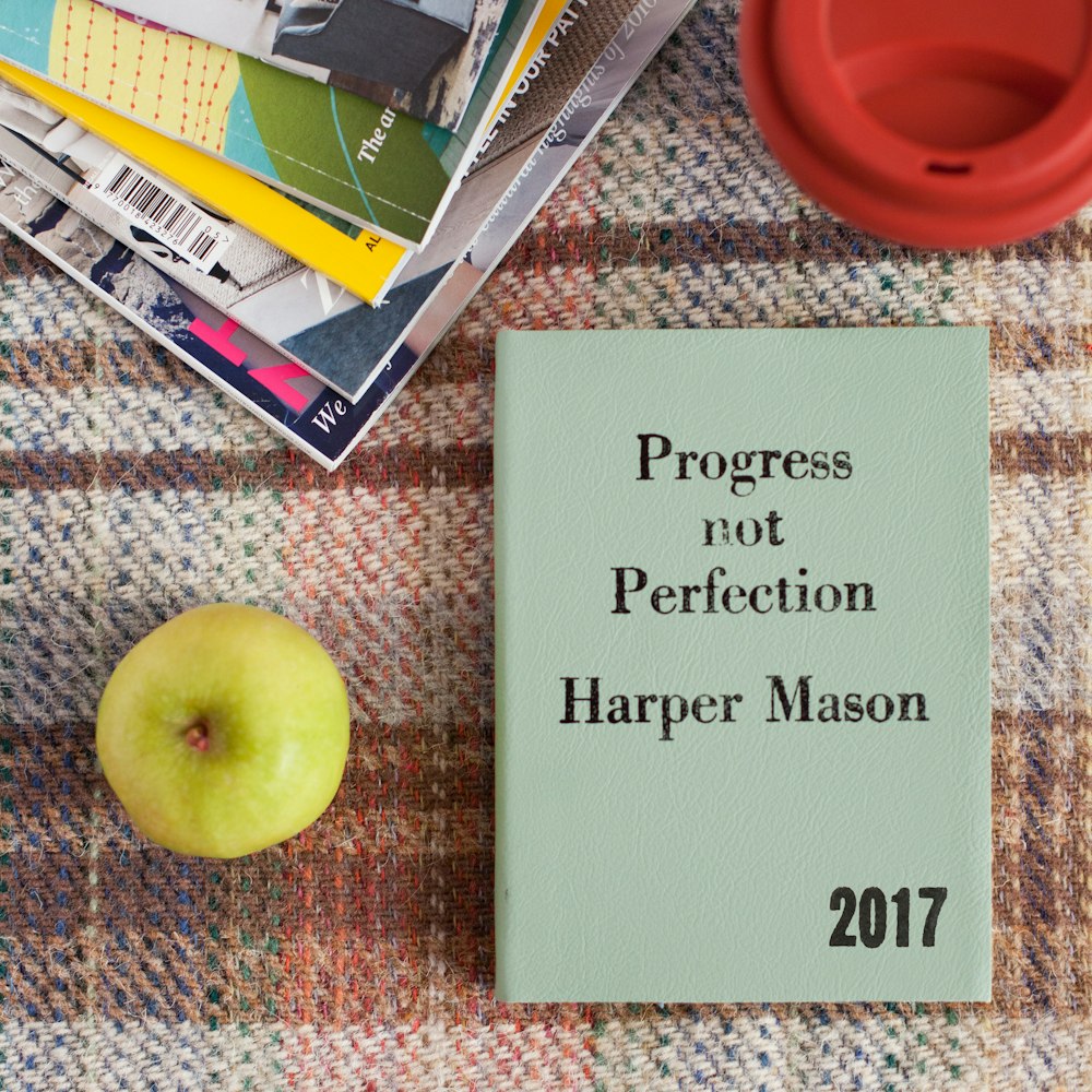 A book that says "Progress not Perfection," written by Harper Mason (2017), placed next to an apple and other books.