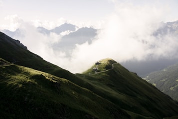 A picturesque mountain landscape featuring lush green hills and a winding path leading to a small building at the crest of a hill. The background showcases towering mountains partially veiled by clouds, creating a serene and majestic atmosphere.