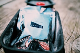 A shiny, reflective package labeled 'LANDER' rests in a black, cushioned tray on a wooden surface. The packaging has a metallic appearance with a mix of colors, including blue and silver tones, and the background is slightly blurred, emphasizing the package.