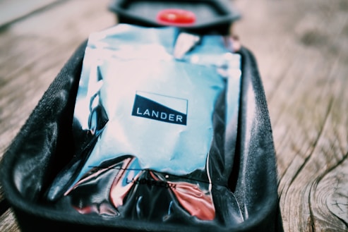 A shiny, reflective package labeled 'LANDER' rests in a black, cushioned tray on a wooden surface. The packaging has a metallic appearance with a mix of colors, including blue and silver tones, and the background is slightly blurred, emphasizing the package.