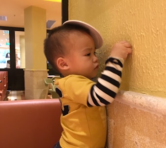 A young child is standing by a textured yellow wall, wearing a yellow shirt with black stripes on the sleeves and a partially worn light-colored cap. The child is focused and appears to be interacting with the wall, possibly trying to touch or examine something.