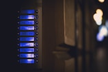A close-up view of an illuminated apartment intercom system with multiple buttons and labels, each emitting a soft blue glow. The surrounding environment appears dimly lit, creating a contrast between the bright interface and the dark background.