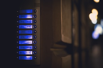 A close-up view of an illuminated apartment intercom system with multiple buttons and labels, each emitting a soft blue glow. The surrounding environment appears dimly lit, creating a contrast between the bright interface and the dark background.