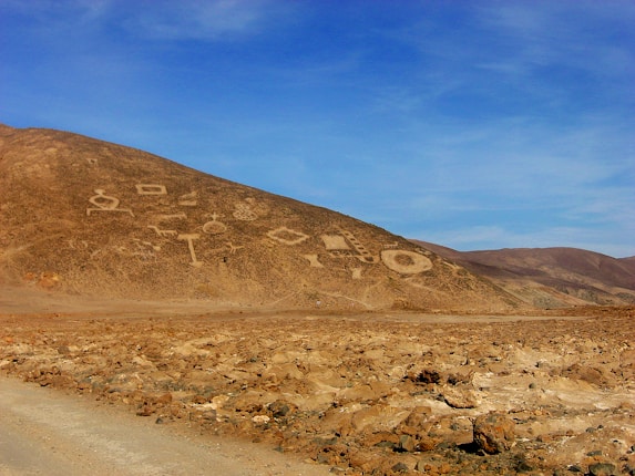 A barren hillside adorned with a series of large geometric geoglyphs, including circles, straight lines, and other angular shapes. The landscape is characterized by a dry, rocky terrain with a clear blue sky overhead.