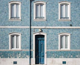 A facade of a building features blue patterned tiles with white framed windows and a large ornate blue door. The wall is adorned with decorative tiles that add texture and character. A vertical pipe runs down the center of the facade, and there are electrical wires above the door.