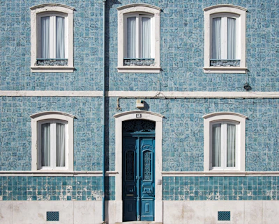 A facade of a building features blue patterned tiles with white framed windows and a large ornate blue door. The wall is adorned with decorative tiles that add texture and character. A vertical pipe runs down the center of the facade, and there are electrical wires above the door.