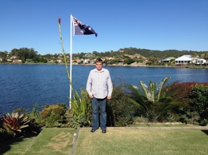 A man stands on a grassy area near a body of water, with lush greenery and a tall plant beside him. An Australian flag is flying on a flagpole, and the background features a lake with houses and hills under a clear blue sky.