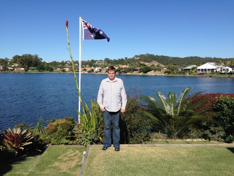 A man stands on a grassy area near a body of water, with lush greenery and a tall plant beside him. An Australian flag is flying on a flagpole, and the background features a lake with houses and hills under a clear blue sky.