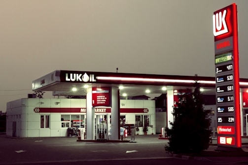 A Lukoil gas station with a modern design features prominently, displaying fuel prices on a tall, illuminated sign. The station includes a minimarket and several fuel pumps under a canopy. The lighting suggests a dusk or dawn setting, casting a soft ambient glow.