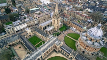 Aerial view of a historic, university campus featuring a domed building and a church with a tall spire surrounded by quadrangles and traditional stone buildings. The architecture is Gothic with intricate details, and the area is interspersed with green lawns and walkways.