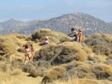 A group of children are playing among dry, bushy vegetation in a sandy and desert-like environment. In the background, arid hills or mountains rise under a hazy sky. The children appear to be exploring or play-acting with sticks, suggesting imaginative play.