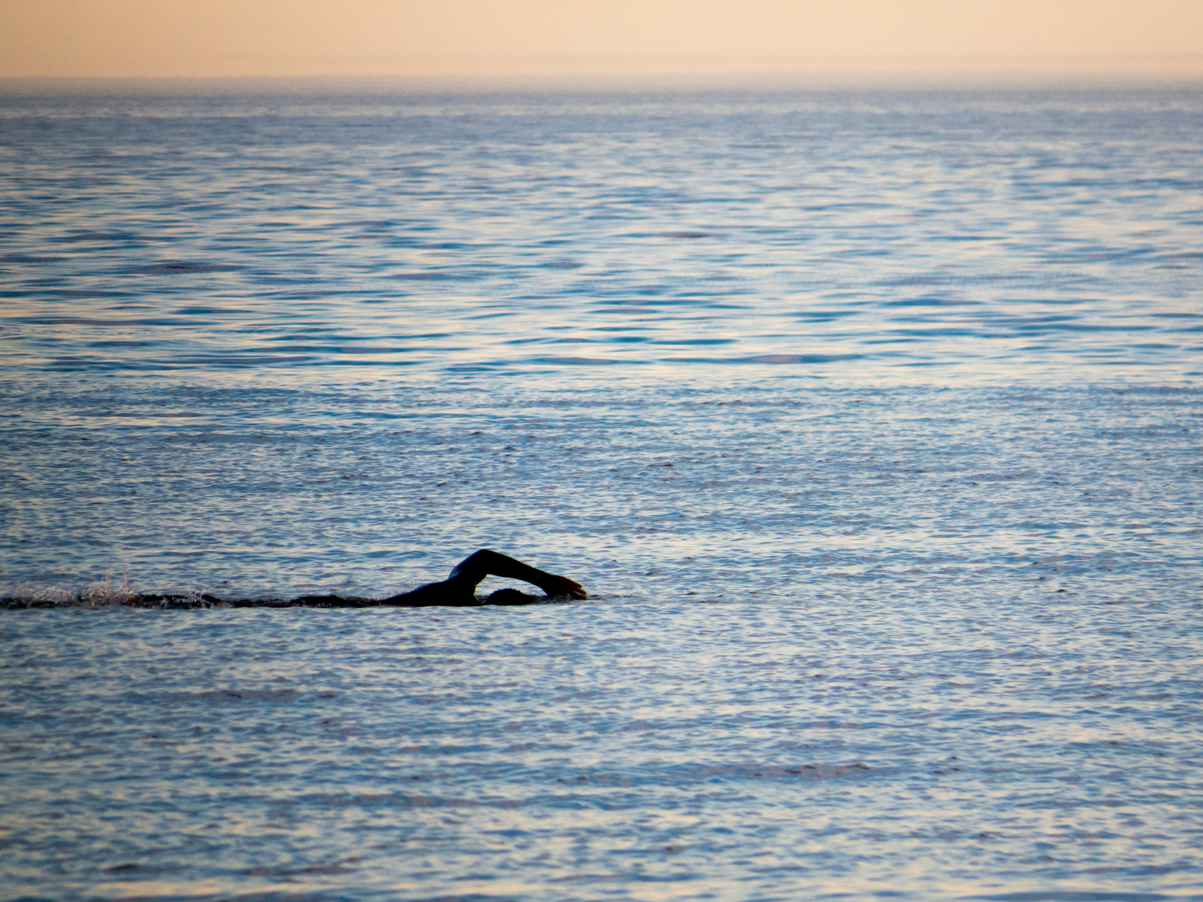 A lone swimmer performs a freestyle stroke in the middle of a calm, vast body of water under a gentle, warm evening sky. The horizon blends softly, suggesting a tranquil and serene environment.