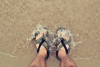 A pair of stylish men's sandals on a sandy beach with waves in the background.