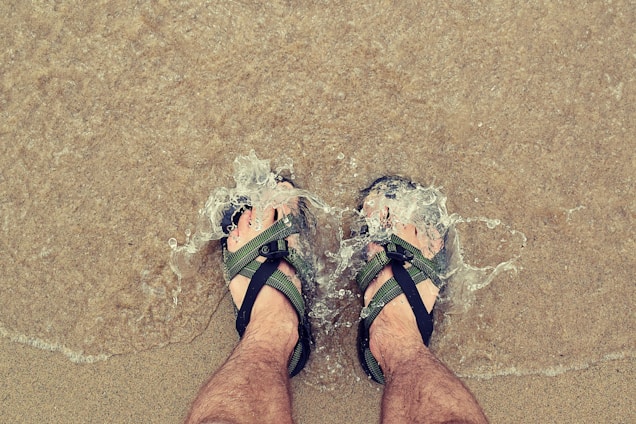 A pair of stylish men's sandals on a sandy beach with waves in the background.