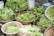 Various types of fresh green vegetables are displayed in woven baskets at a market. The leafy greens and long beans are arranged neatly, showcasing their freshness and vibrant colors.