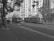 A street scene featuring an old-fashioned streetcar and a bus traveling along tracks and a road adjacent to tall, ornate buildings. Trees line the sidewalk, and overhead wiring indicates electric transit in use. The architecture suggests a historic urban area.