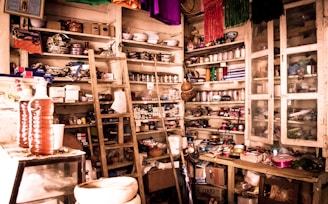 A cluttered room filled with wooden shelves packed with various items such as jars, boxes, and household goods. There is a wooden ladder leaning against the shelves, and multiple colorful fabrics hang from the ceiling. The room appears to be used for storage, displaying a mix of household items and products.