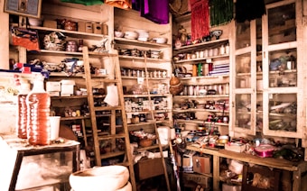 A cluttered room filled with wooden shelves packed with various items such as jars, boxes, and household goods. There is a wooden ladder leaning against the shelves, and multiple colorful fabrics hang from the ceiling. The room appears to be used for storage, displaying a mix of household items and products.