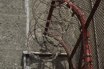 A metal fence with coiled razor wire running along its top, casting shadows on a concrete surface. The fence has a red support structure adding a touch of color to the otherwise muted scene.
