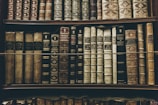 A collection of vintage, leather-bound books arranged neatly on wooden shelves. The spines of the books display intricate gold embossing and titles, showcasing a sense of antiquity and elegance.
