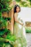 A joyful woman in a softly flowing terracotta-toned dress standing against a cream ivory wall, sunlight casting a warm glow.