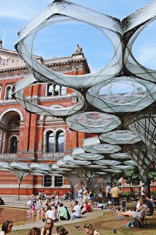 A group of people are gathered in an outdoor area next to a large historic building made of red brick. Above them, a series of interconnected net-like structures are suspended, creating a unique installation or art piece. Some people are sitting on the ground, while others are standing and interacting with one another. The scene is set in a courtyard with arches and detailed architectural features on the building.