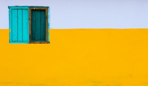 A bright, colorful wall featuring a bold horizontal division of colors with the upper part painted white and the lower portion painted yellow. On the left side, a simple, rustic window with blue-green wooden shutters adds a contrasting pop of color.