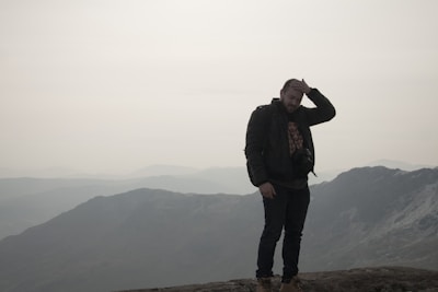 A person stands on a rocky peak, looking contemplative with one hand on their head. The backdrop features misty mountains under a cloudy sky, creating a subdued and introspective atmosphere.